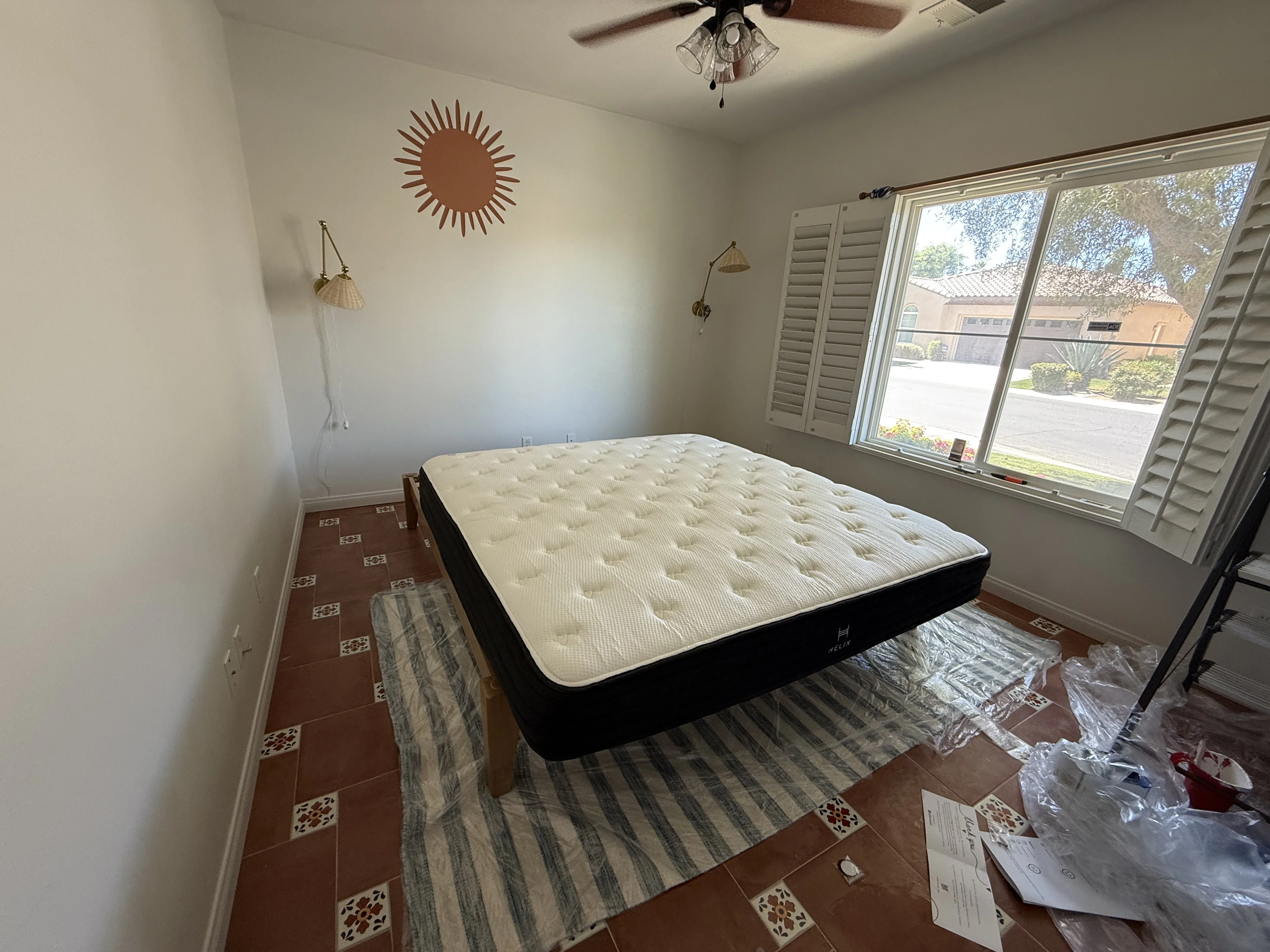 King mattress delivered to the Cali Room at Terra Luz during renovation — Helix mattress on the floor with blue striped jute rug, no headboard yet, brass sconces and sun wall art on white wall