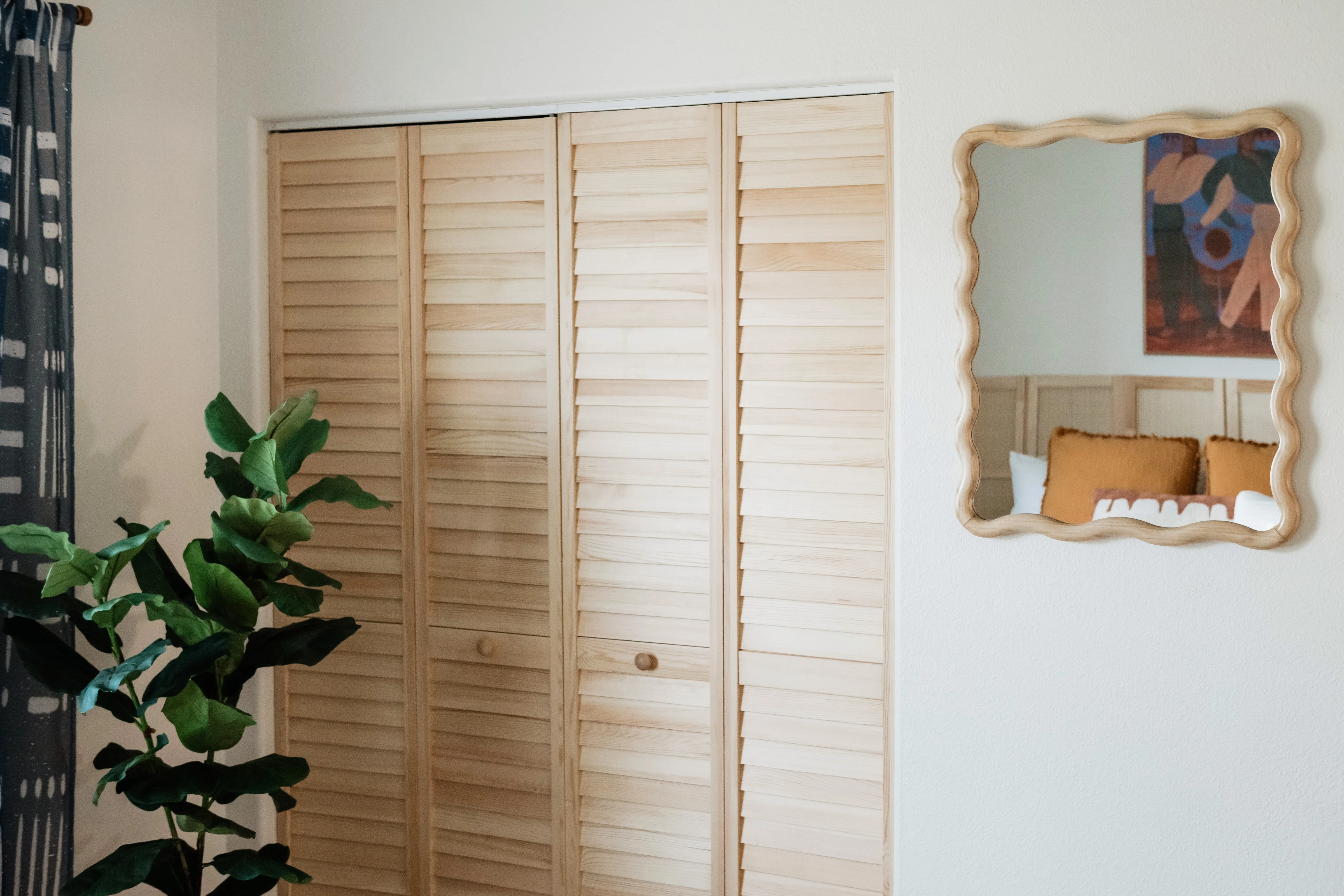 Stained natural louvered bifold closet doors and wavy wood mirror in the Cali Room at Terra Luz, with fiddle leaf fig and mudcloth curtains