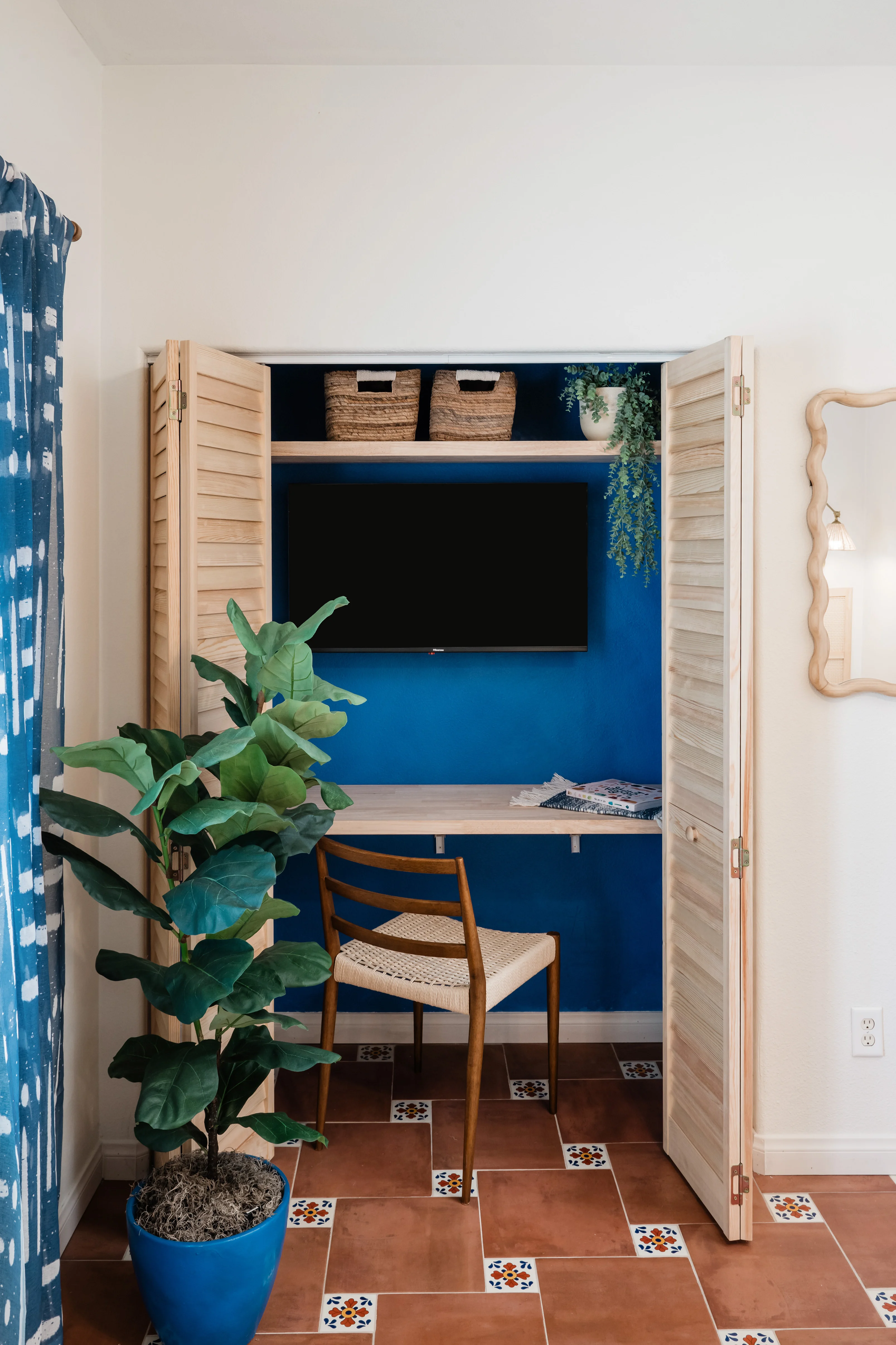 Louvered bifold doors fully open revealing the Frida Kahlo blue workstation in the Cali Room at Terra Luz, with floating desk, rattan chair, woven storage baskets, and blue fiddle leaf fig pot