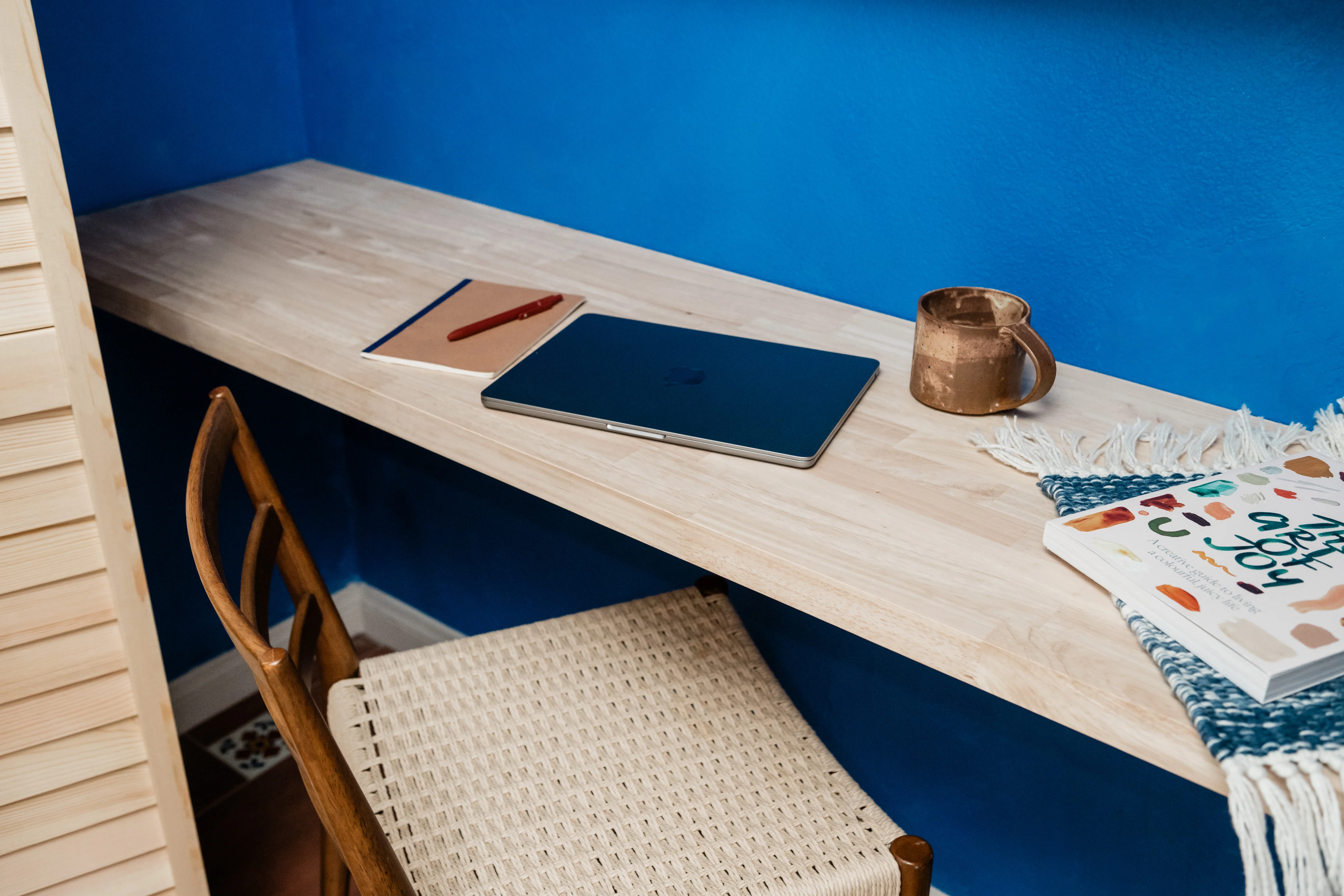 Kahlo Blue lime-washed workstation wall with floating live-edge desk, rattan wishbone chair, and handmade ceramic mug in the Cali Room at Terra Luz