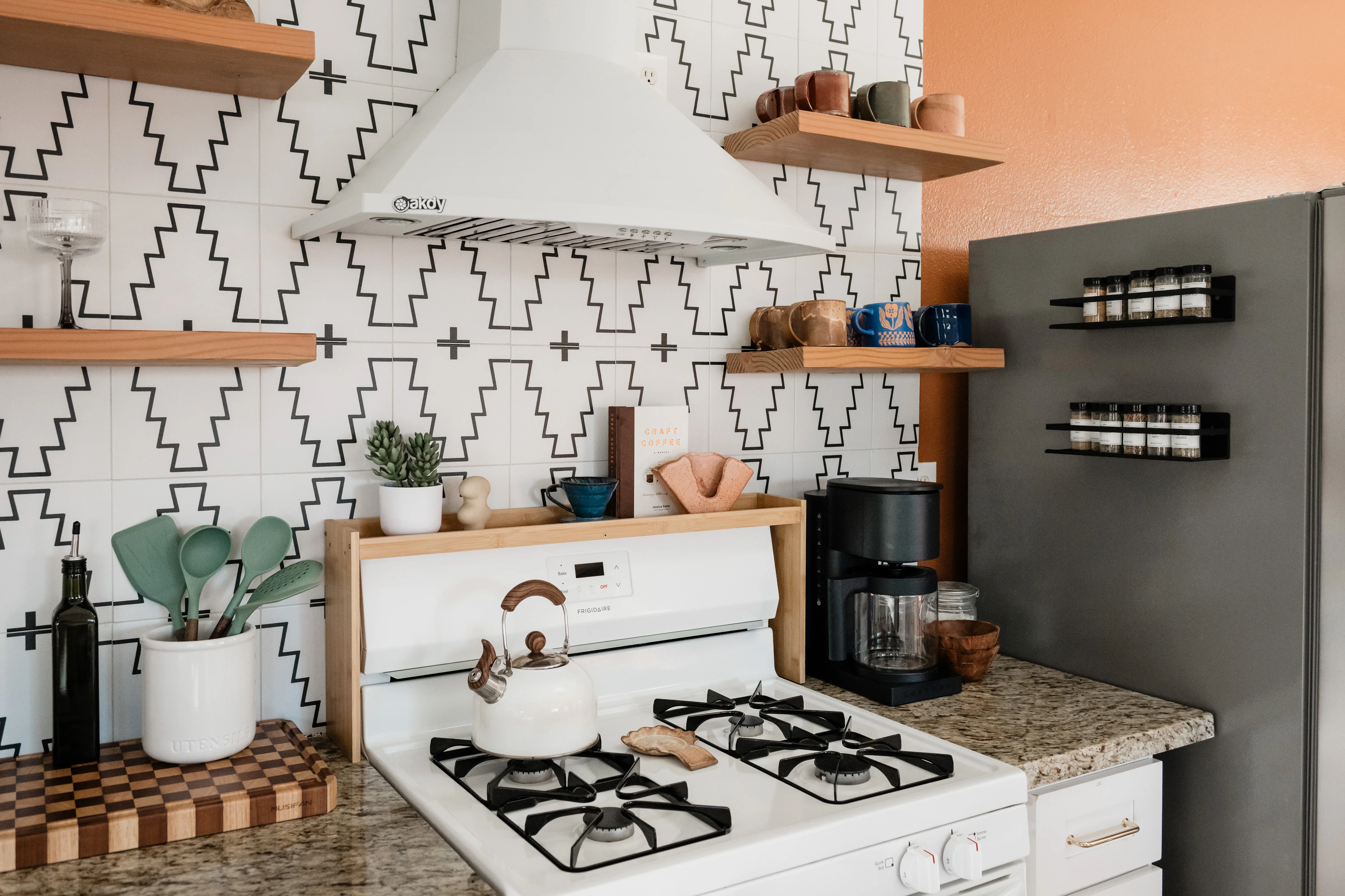 Terra Luz kitchen with white gas range, graphic black and white tile backsplash, open wood shelving with ceramics and glassware, and coffee station at left