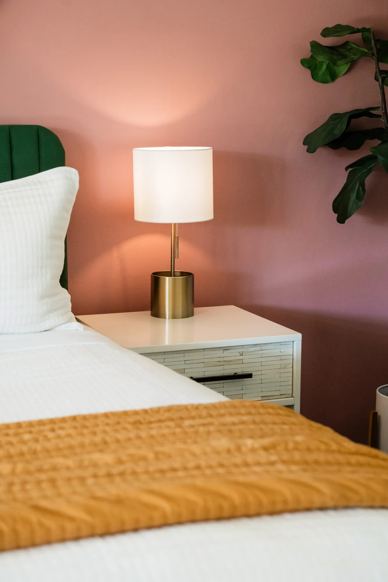 Cozy Cactus primary bedroom nightstand with gold lamp, pink accent wall, and fiddle leaf fig