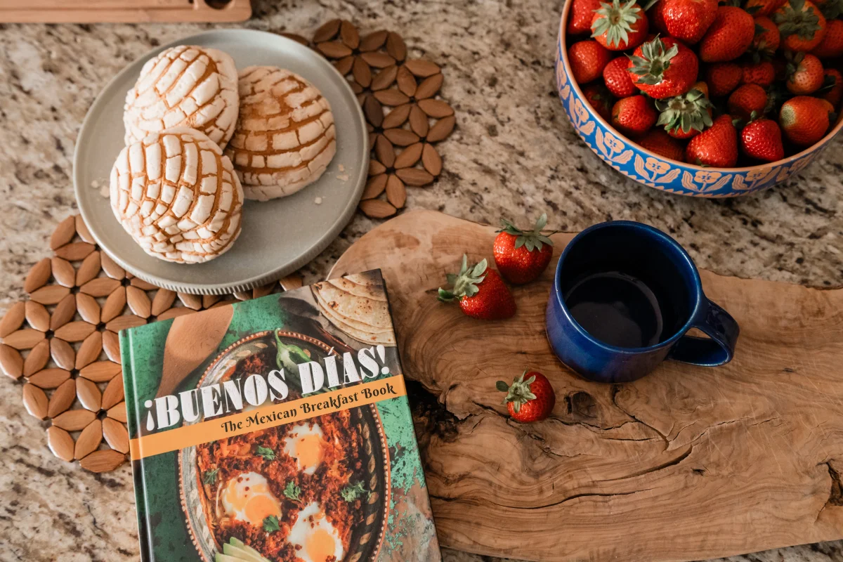 Terra Luz kitchen counter with Buenos Dias cookbook, strawberries, pan dulce, and blue mug on acacia board