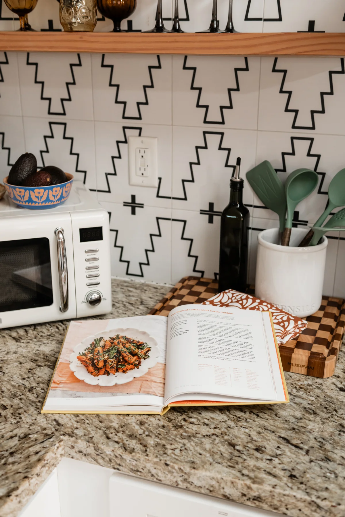 Terra Luz kitchen with open cookbook on granite counter, geometric backsplash, retro microwave, and kitchen tools