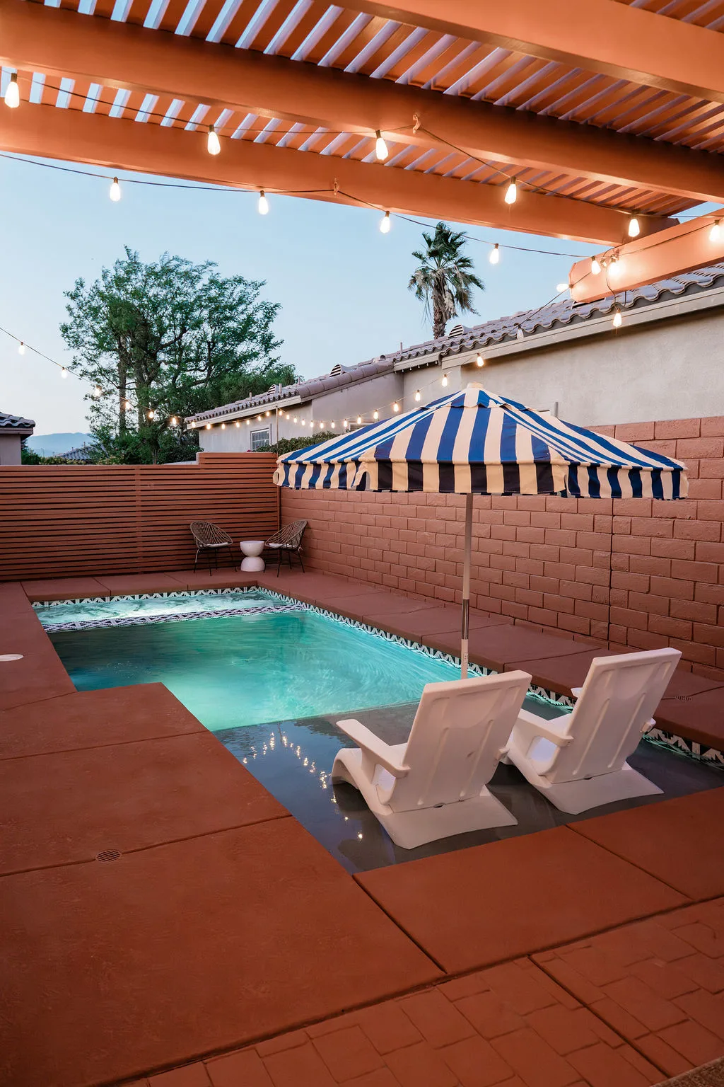 Terra Luz pool at dusk with string lights, two white Adirondack chairs, and blue white umbrella