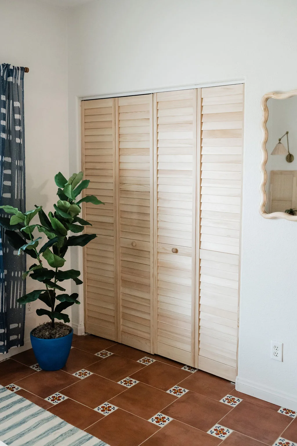 Terra Luz Cali bedroom louvered closet doors with mudcloth curtains, blue striped rug, Mexican tile floor, fiddle leaf fig in blue pot, and wavy mirror with wall sconce