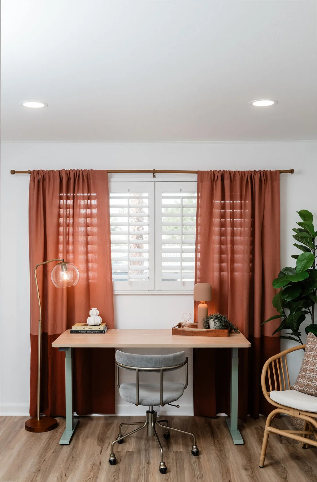 The Sundune Palm Springs second bedroom with rust linen curtains, standing desk, rattan chair, and natural light