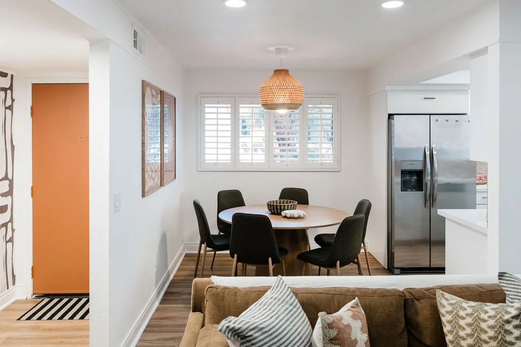 The Sundune Palm Springs dining area with round walnut table, olive green chairs, wicker pendant, and kitchen beyond